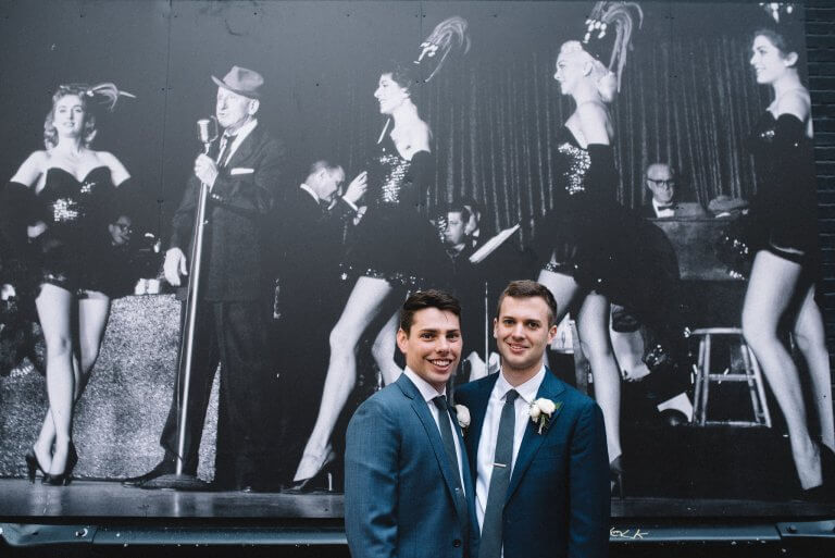grooms in blue suits smile in front of chez mural