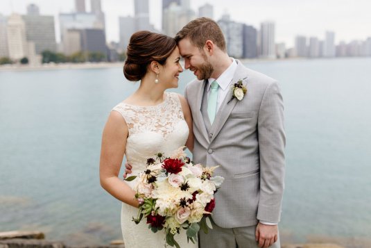 bride in white lace dress and groom in grey suit pose in front of chicago lake