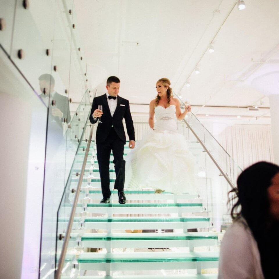 bride and groom walk down glass staircase with champagne in hand