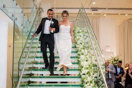 bride and groom enter down glass stair case lined with white and green flowers