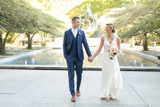 bride and grrom in blue suit hold hands at art institute fountain